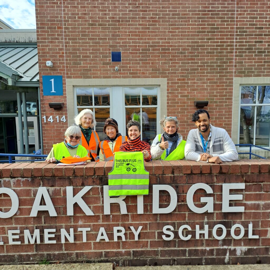 Oakridge Elementary School sign. People standing behind holding a vest that says this bus is us.