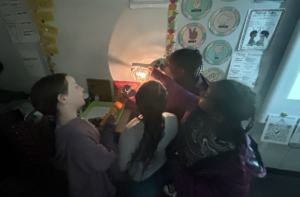 4 students working together to shine light, with a flash light, onto tin foil to see it reflect and leave a shadow.