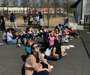 Ms. Fitzpatrick's class sitting outside on the black top with their solar eclipse glasses on, observing the solar eclipse.