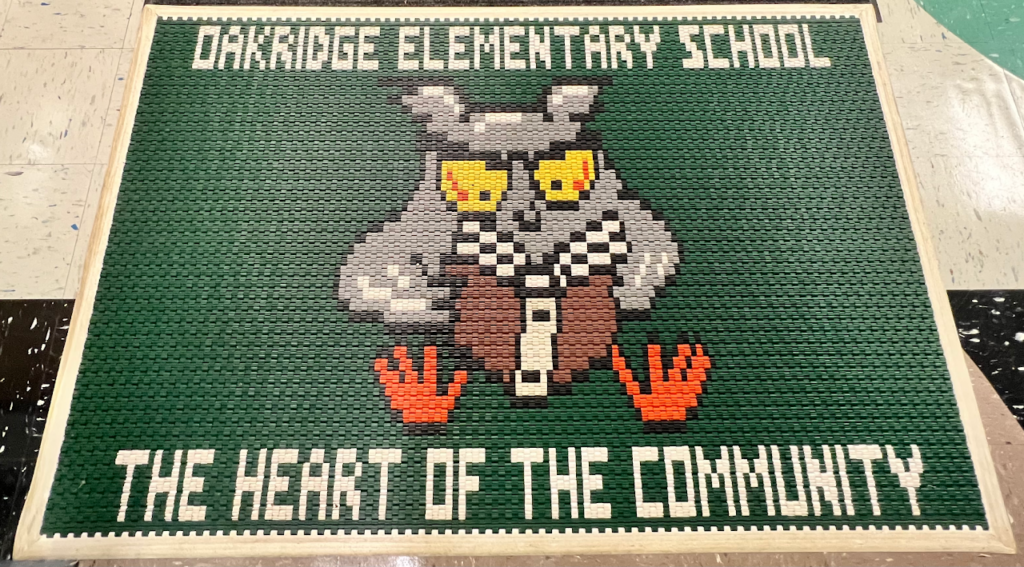 A picture of the welcome mat at the school. It is green with an owl in the middle reading a book. The top says Oakridge Elementary School and the bottom says The Heart of the Community.