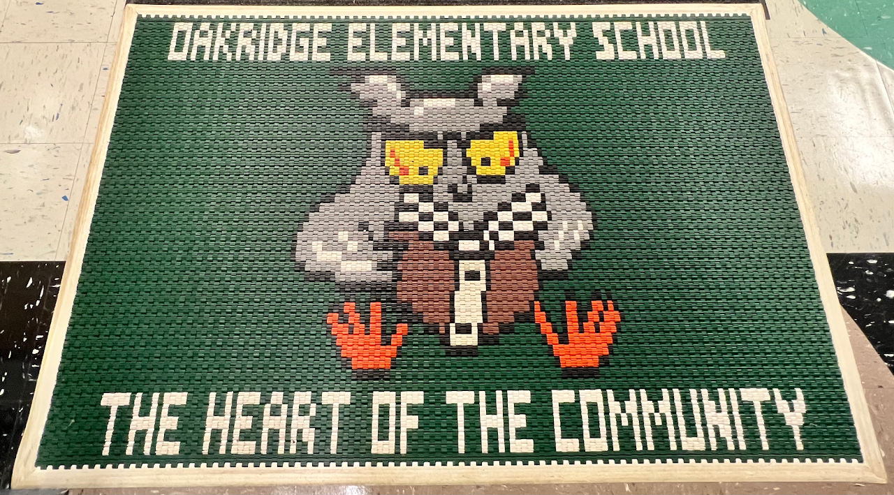 A picture of the welcome mat at the school. It is green with an owl in the middle reading a book. The top says Oakridge Elementary School and the bottom says The Heart of the Community.